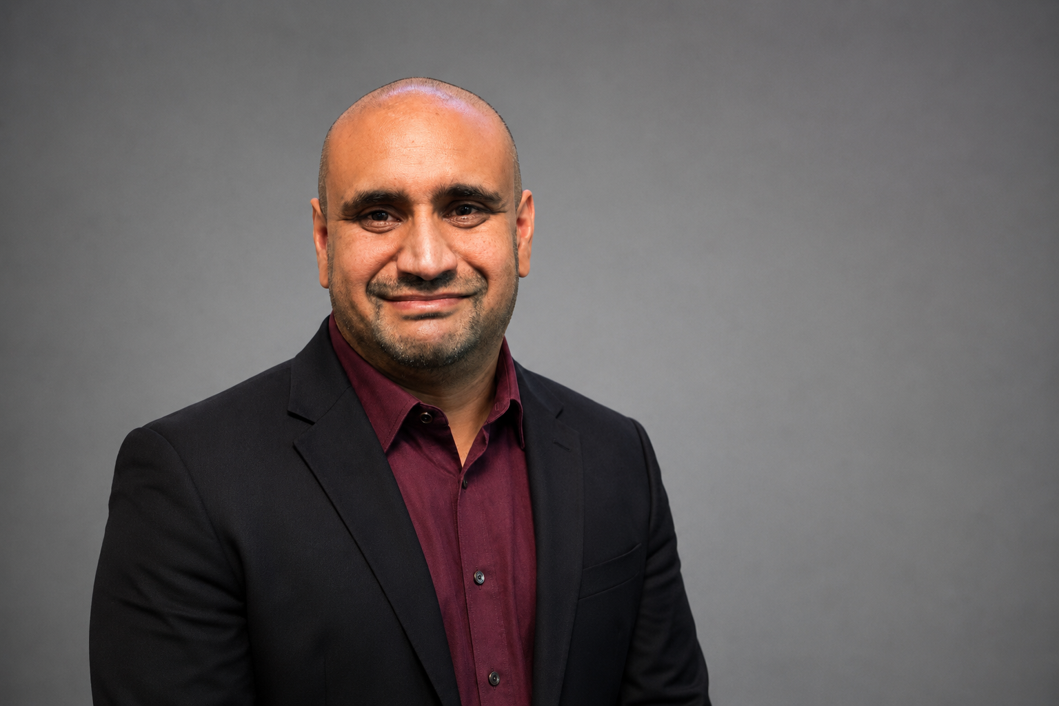 A man with a shaved head and beard, dressed in a dark blazer and a maroon shirt, smiling at the camera against a plain gray background.