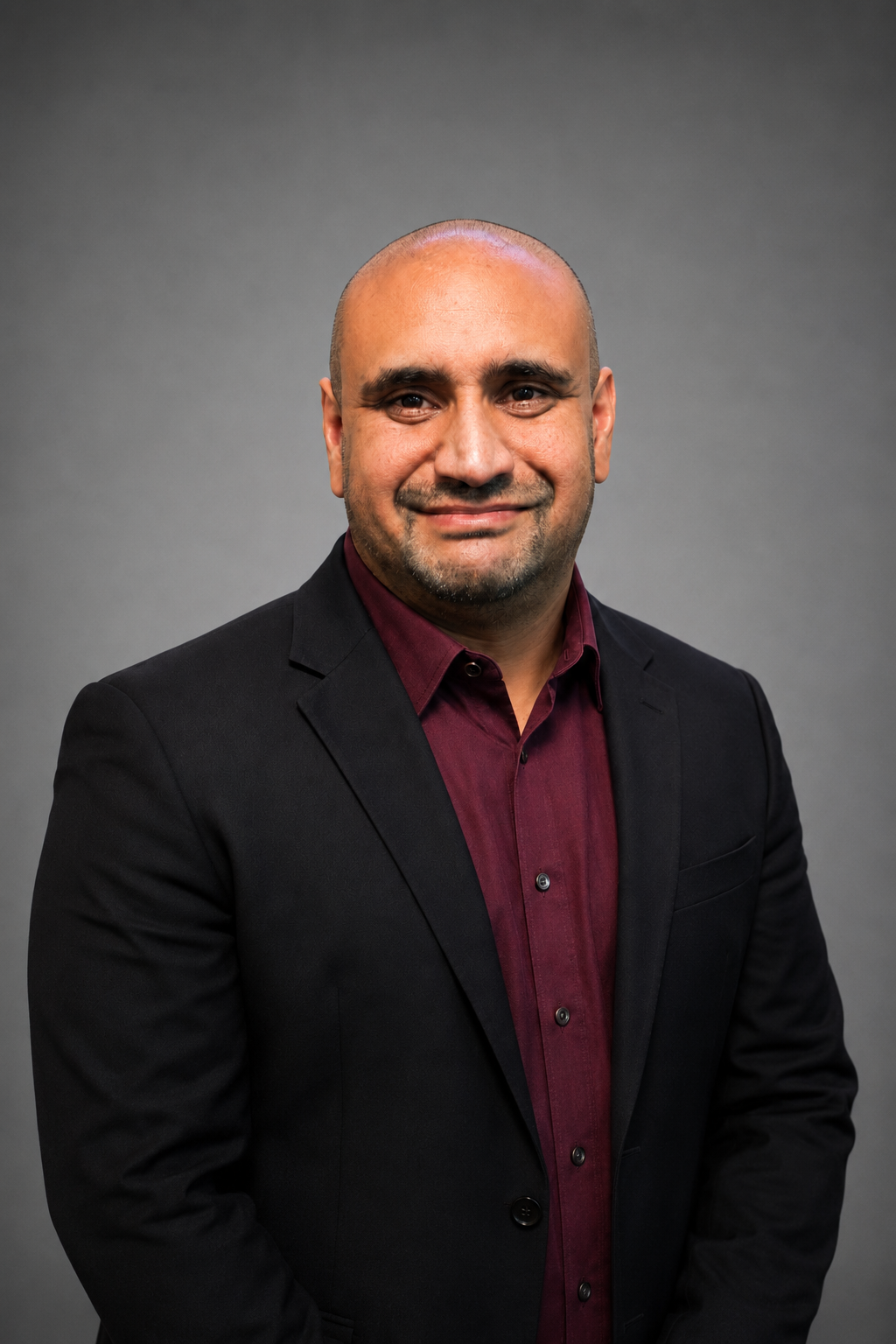 A man in a black suit and maroon shirt smiling at the camera against a gray background.