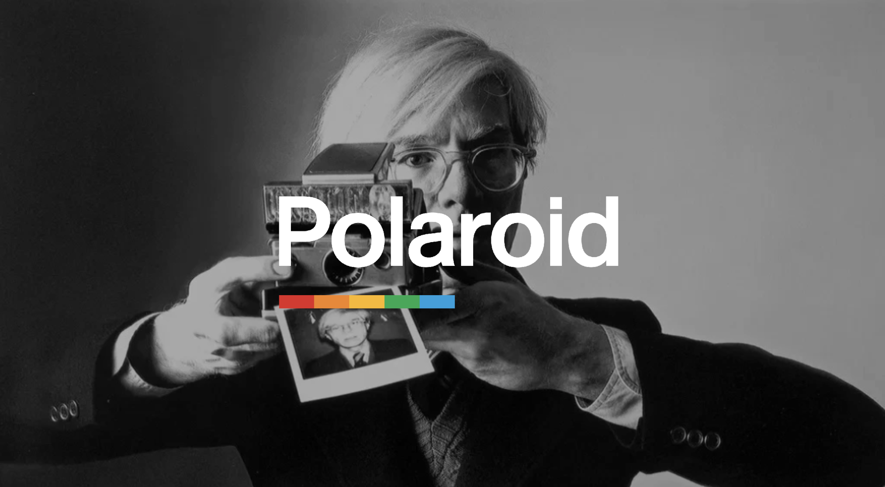 A person with light-colored hair and glasses holding a vintage camera with a Polaroid picture, black and white photo