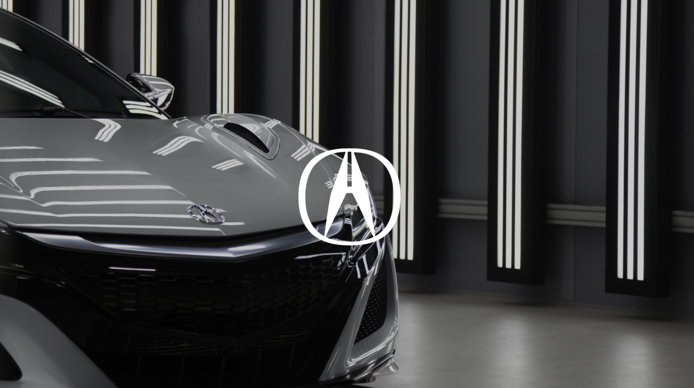 Close-up of the front portion of a silver sports car with an Acura logo, parked indoors with a modern black background and vertical light strips.
