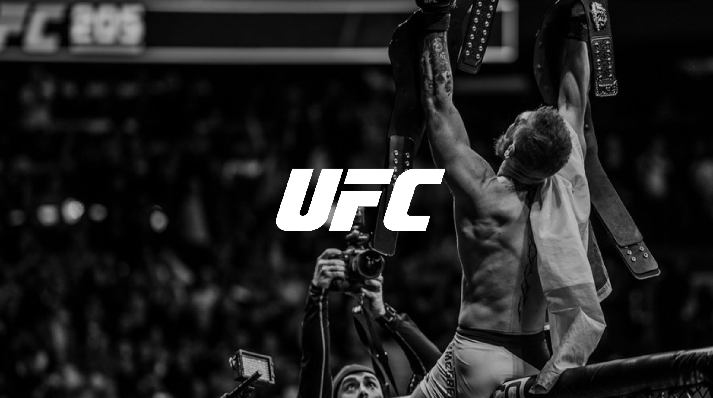 A mixed martial arts fighter holding a championship belt above his head after a victory in the UFC, with a photographer capturing the moment and a crowd in the background.