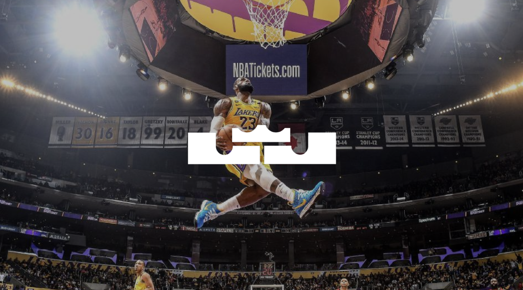 NBA player in Los Angeles Lakers uniform jumping with a basketball in front of a basketball hoop in a large indoor arena filled with spectators and banners.