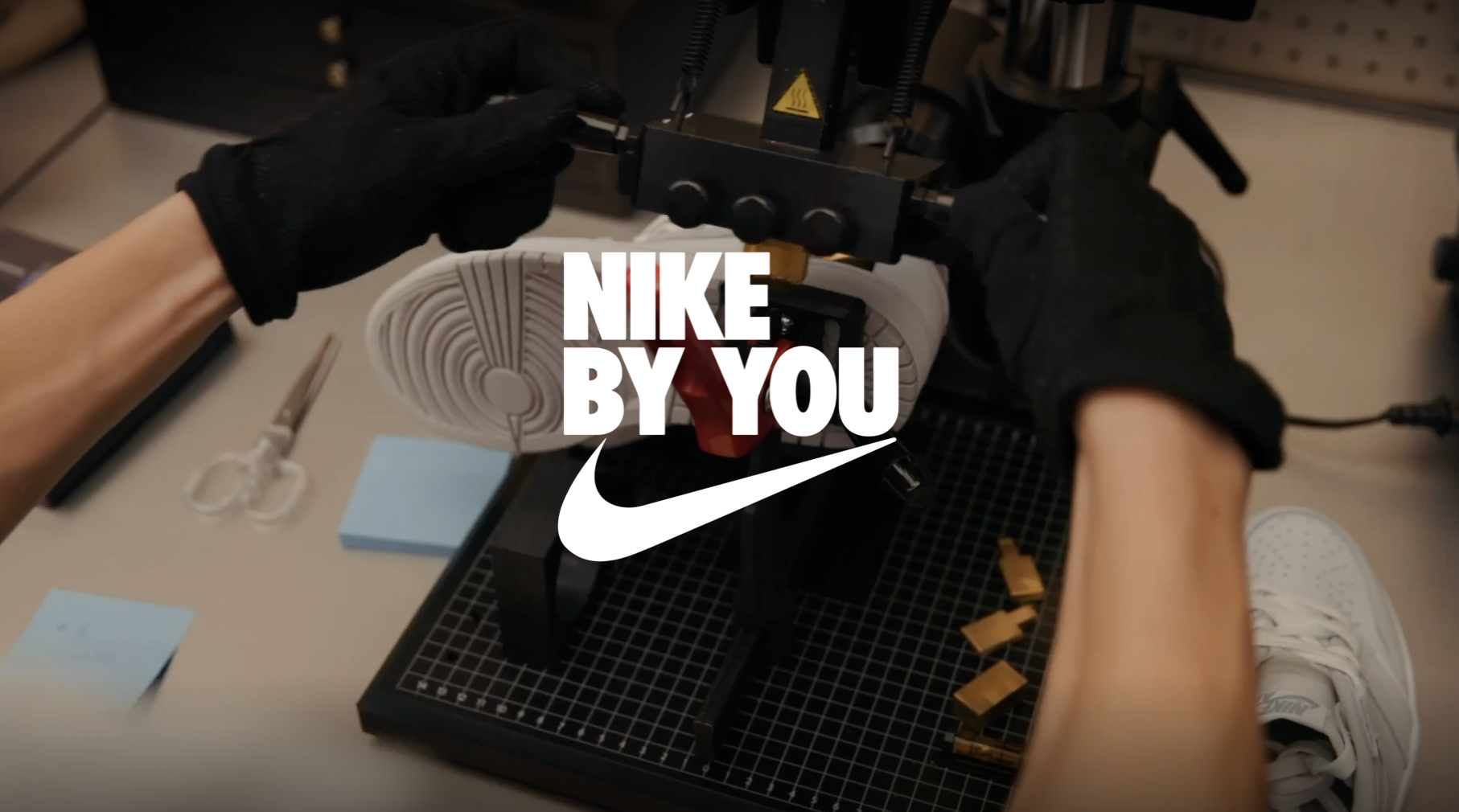 A pair of hands in black gloves assembling a white Nike sneaker with a grid workspace, scissors, sticky notes, and small orange components nearby, overlaid with the Nike logo and the text 'NIKE BY YOU'.
