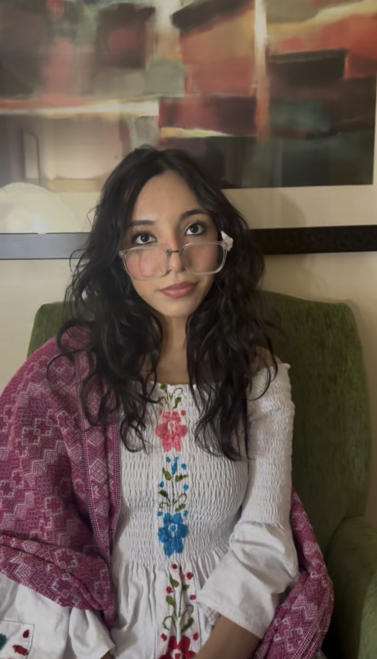 A woman with dark, wavy hair, glasses, and a white embroidered dress, with a purple rebozo draped on her shoulders, sitting on a green chair in front of an abstract painting.