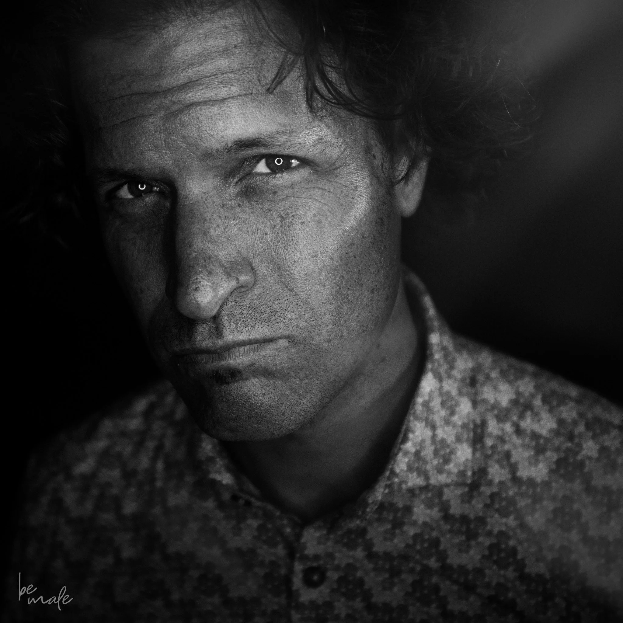 Schwarz-weiß-Porträt eines Mannes mit wilden Haaren, der direkt in die Kamera schaut, mit detaillierter Hautstruktur und einem nachdenklichen Ausdruck.