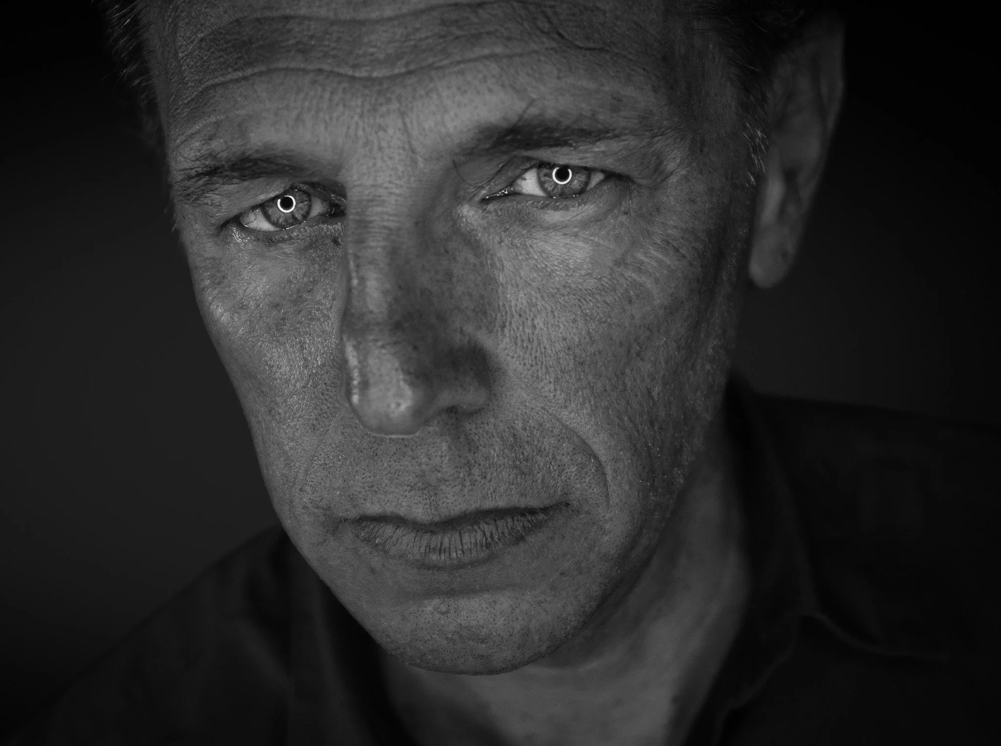 Schwarz-Weiß-Porträt eines Mannes mit intensiven Blick, kurze Haare, auffällige Hautstruktur mit Falten und Textur. Schlichte dunkle Kleidung, dunkler Hintergrund.
