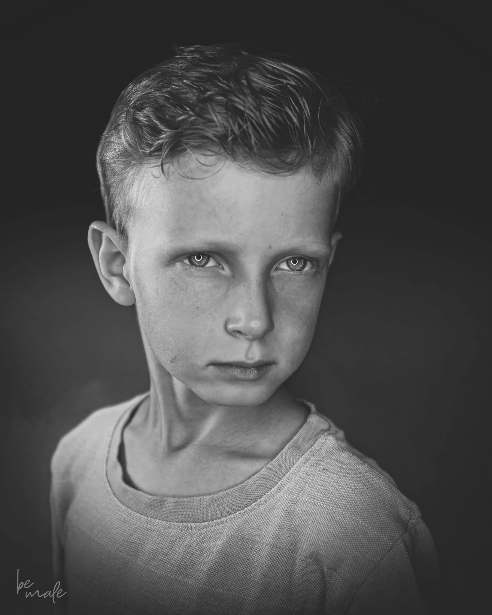 Schwarz-Weiß-Fotografie eines jungen Jungen mit intensiven Blick, kurze Haare, einfaches T-Shirt, dunklem Hintergrund.