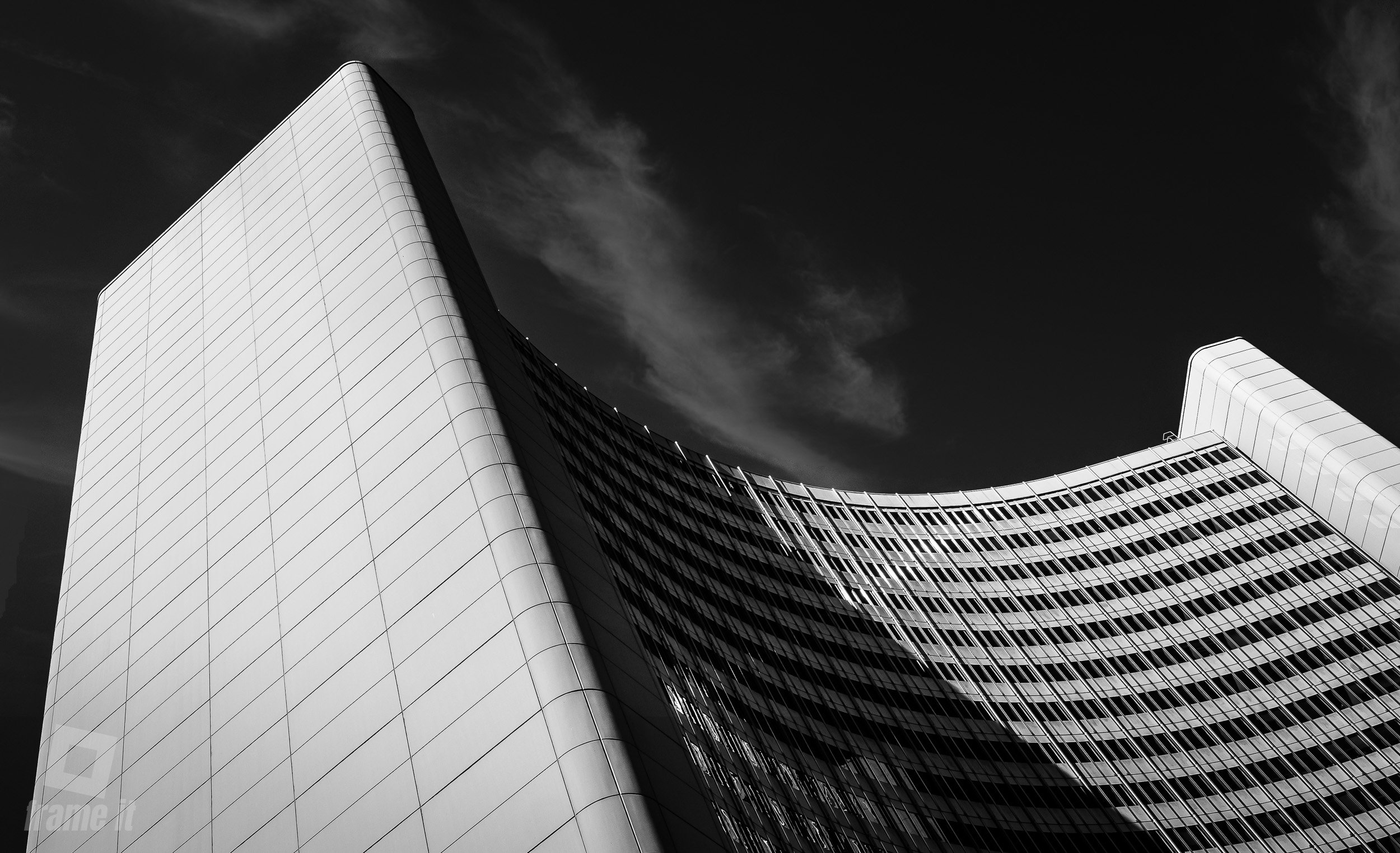 Schwarz-weiße Aufnahme eines modernen, geschwungenen Wolkenkratzers mit Glasfassade gegen einen dunklen Himmel.