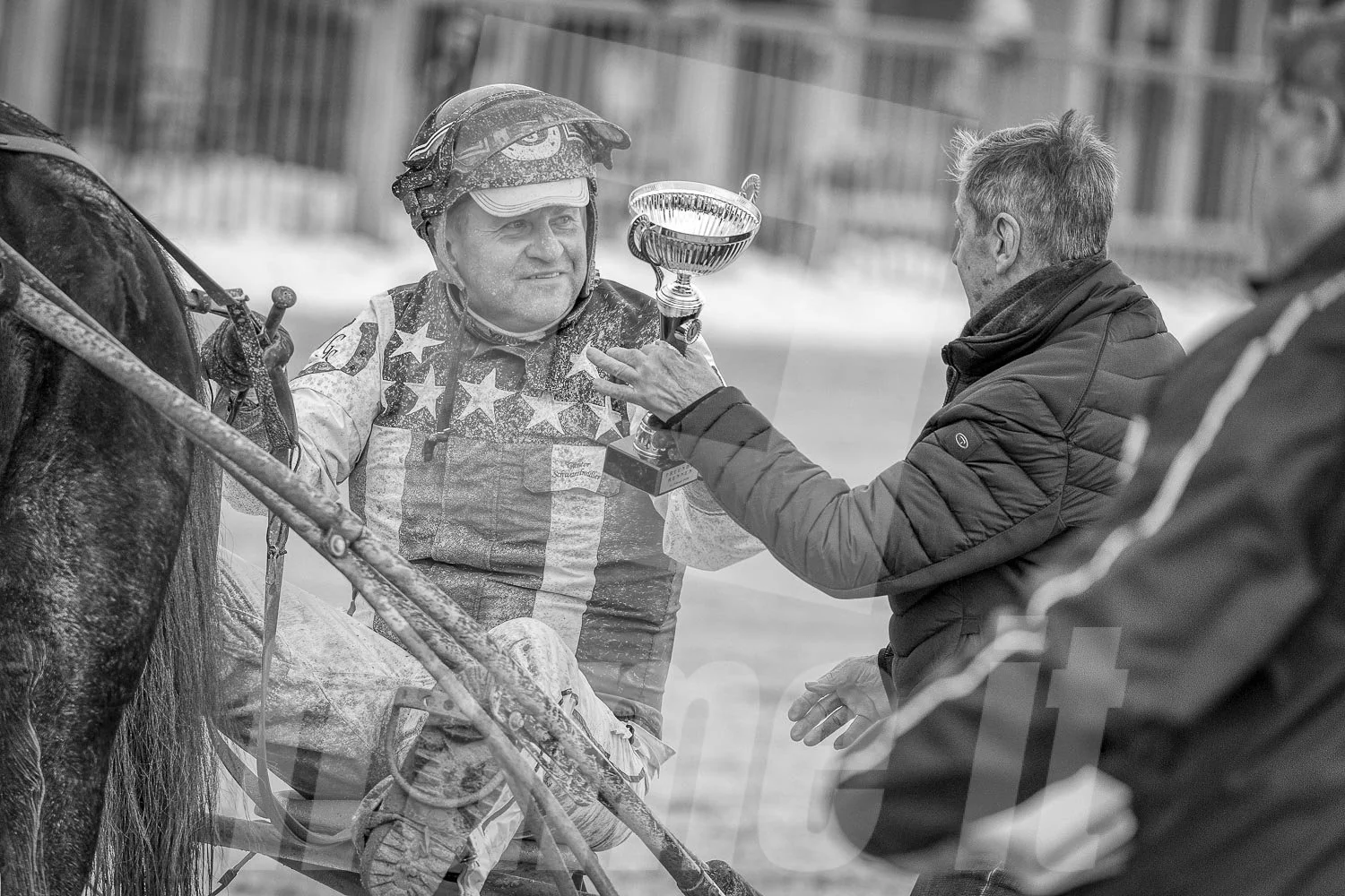 Ein Sieger im Pferderennen erhält eine Trophäe, während ein Mann ihm gratuliert. Der Sieger trägt einen Helm und einen Rennanzug, im Hintergrund sind Zäune sichtbar.