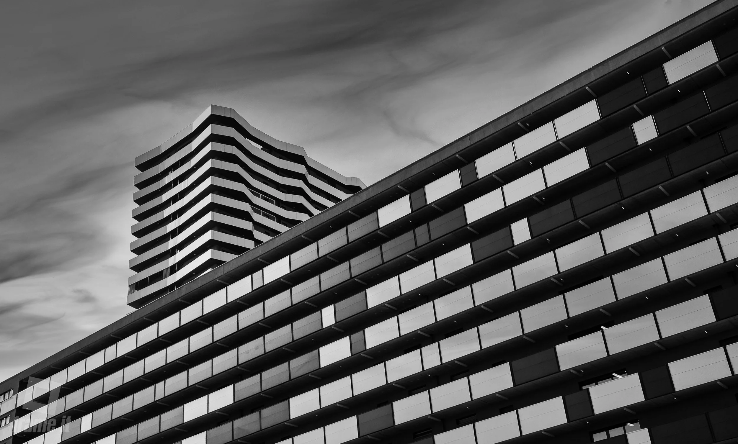 Schwarz-weiß Aufnahme eines modernen Hochhauses mit wellenförmigen Balkonen und klaren Linien gegen einen Himmel mit Wolken, in Wien beim Citygate.