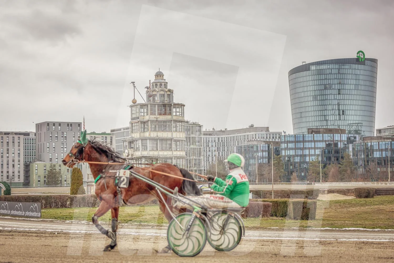 Trabrennpferd mit Trabrennfahrer auf der Pferderennbahn Wien Krieau bei winterlichen Bedingungen, im Hintergrund der historische Richterturm und die modernen Gebäude von der OMV und Bitpanda.