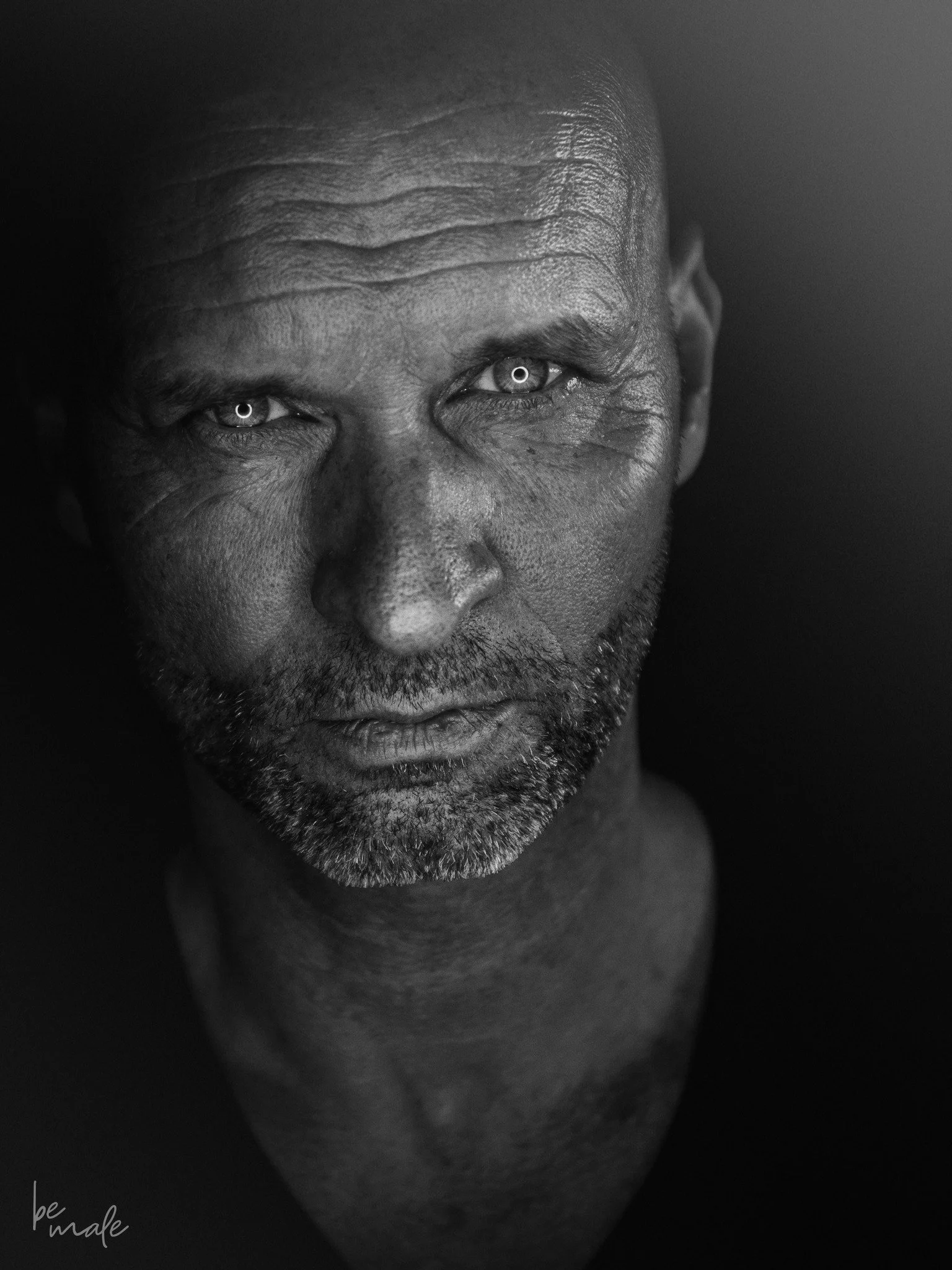 Schwarz-weiß Porträt eines ernst blickenden Mannes mit grauem Bart und kurzen Haaren, fokussiert auf sein Gesicht und seine Augen.