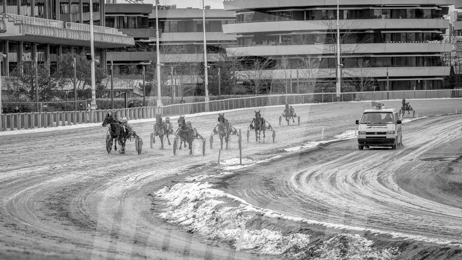 Schwarz-weiss Foto eines Trabrennpferdes, mit einem Trabrennfahrer auf der Trabrennbahn in der Wiener Krieau. Rechts der Wagen der Rennleitung.