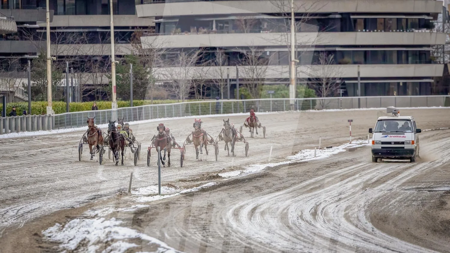 Pferderennen auf einer Sandbahn mit mehreren Pferden und Wagen, ein Rennleitungsfahrzeug fährt auf der rechten Seite, im Hintergrund sind moderne Gebäude und Zuschauer.