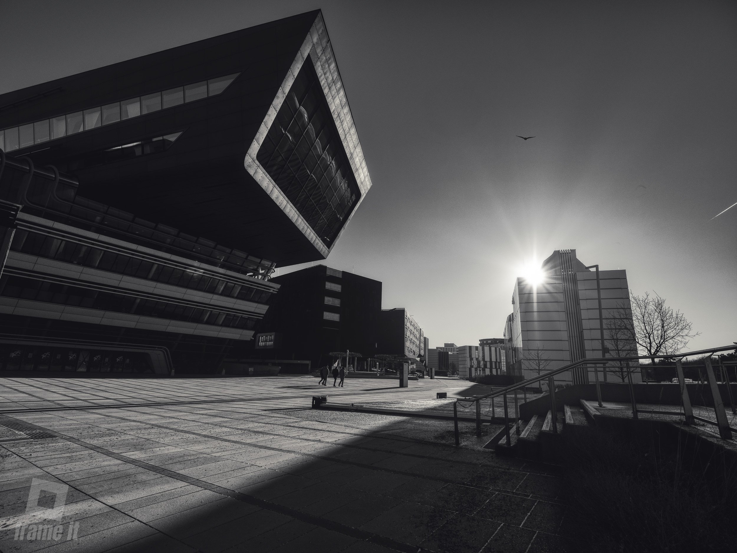 Modernes Gebäude in Wien, die WU, mit Sonnenaufgang im Hintergrund, Schatten auf der Straße, wenige Menschen zu Fuß unterwegs.