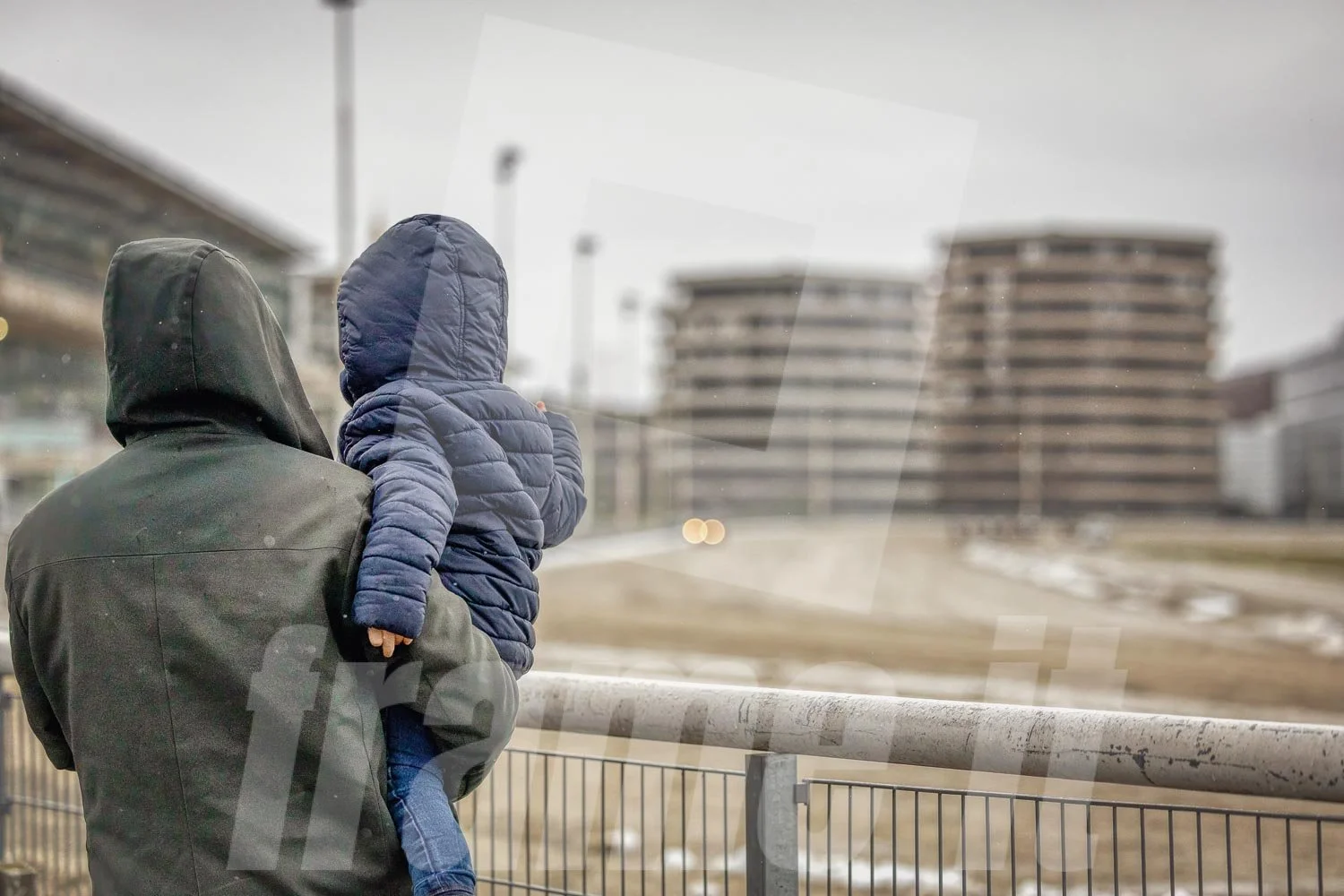 Ein Erwachsener trägt einen schwarzen Kapuzenpullover und hält ein Kind im blauen Jacke. Sie stehen an einem Zaun und schauen auf eine leere Trabrennbahn in der Wiener Krieau.