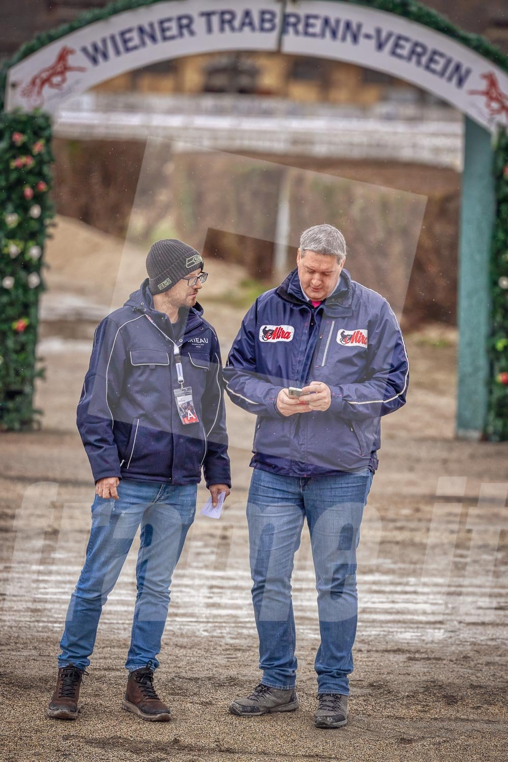 Zwei Männer stehen unter einem Eingangsbogen mit der Aufschrift "Wiener Trabrenn-Verein". Ein Mann schaut auf sein Smartphone, der andere hält ein Stück Papier. Es ist neblig und regnerisch.