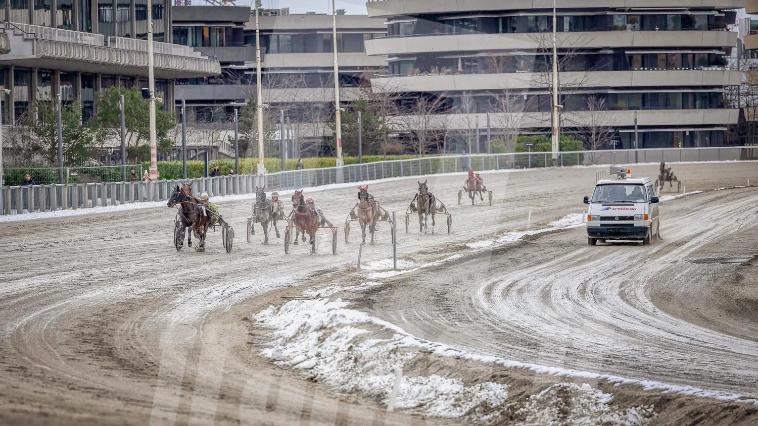 Pferderennen auf einer Wettkampfbahn, mit mehreren laufenden Rennpferden und einem Begleitfahrzeug.