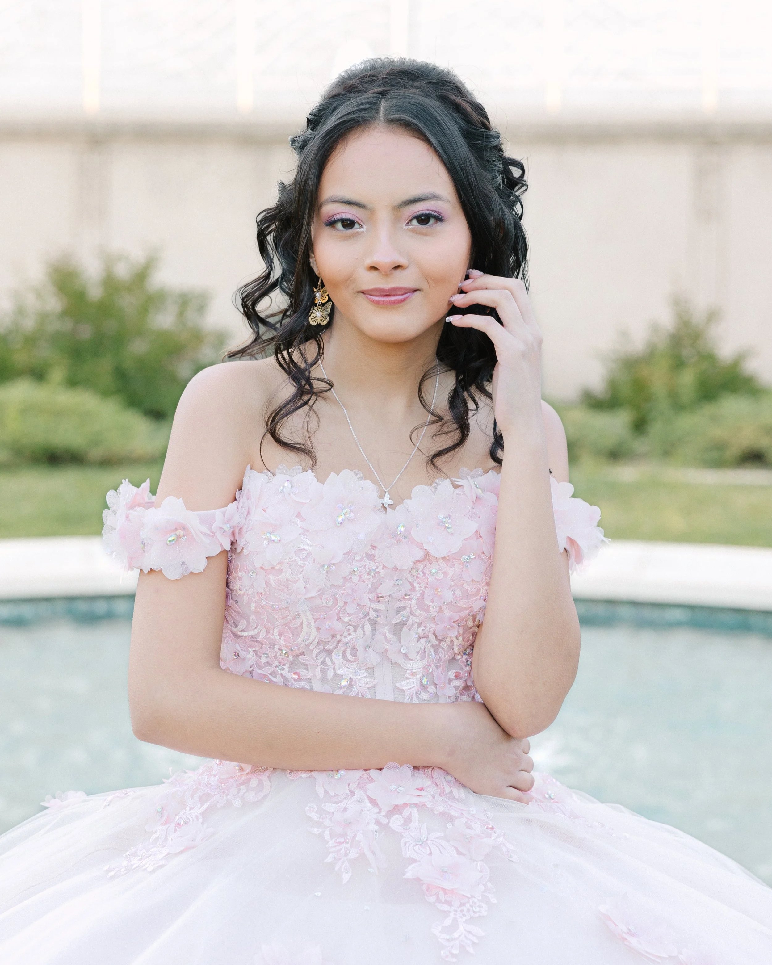 Portrait of a young woman in a pink floral dress with off-the-shoulder sleeves, standing outdoors near a fountain with greenery in the background.