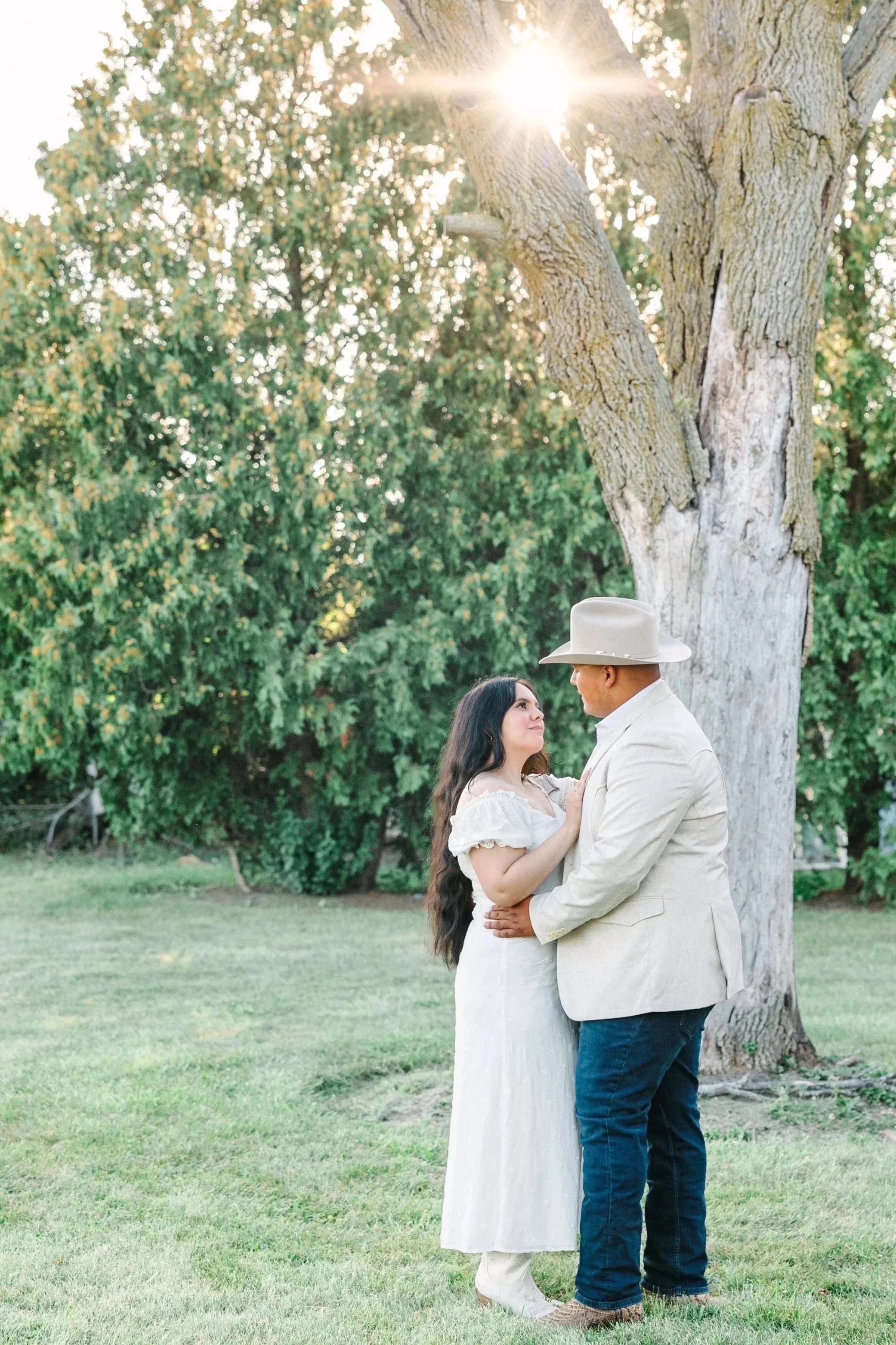A man and woman stand close together under a large tree in a grassy area, with sunlight filtering through the branches. The woman has long dark hair and a white dress, and the man is wearing a beige jacket, dark jeans, and a wide-brimmed hat.