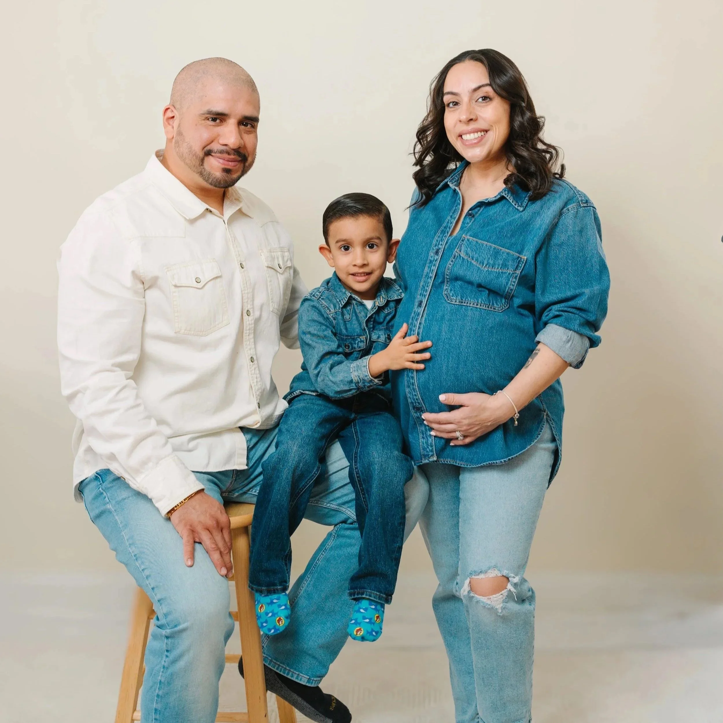 Family of three with a pregnant woman, middle son sitting on her belly, father on stool to her left, all smiling against a plain background.