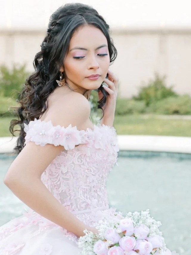 Young woman in a pink, floral, off-shoulder dress holding a bouquet of pink and white roses, with dark wavy hair and earrings, standing outdoors near a pool.