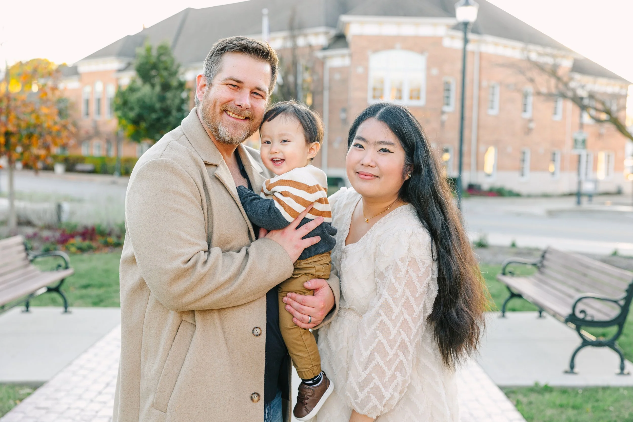 A family of three outdoors in front of a large brick building. The man with a beard and light coat is holding a smiling young boy, and a woman with long dark hair in a light sweater stands beside them.