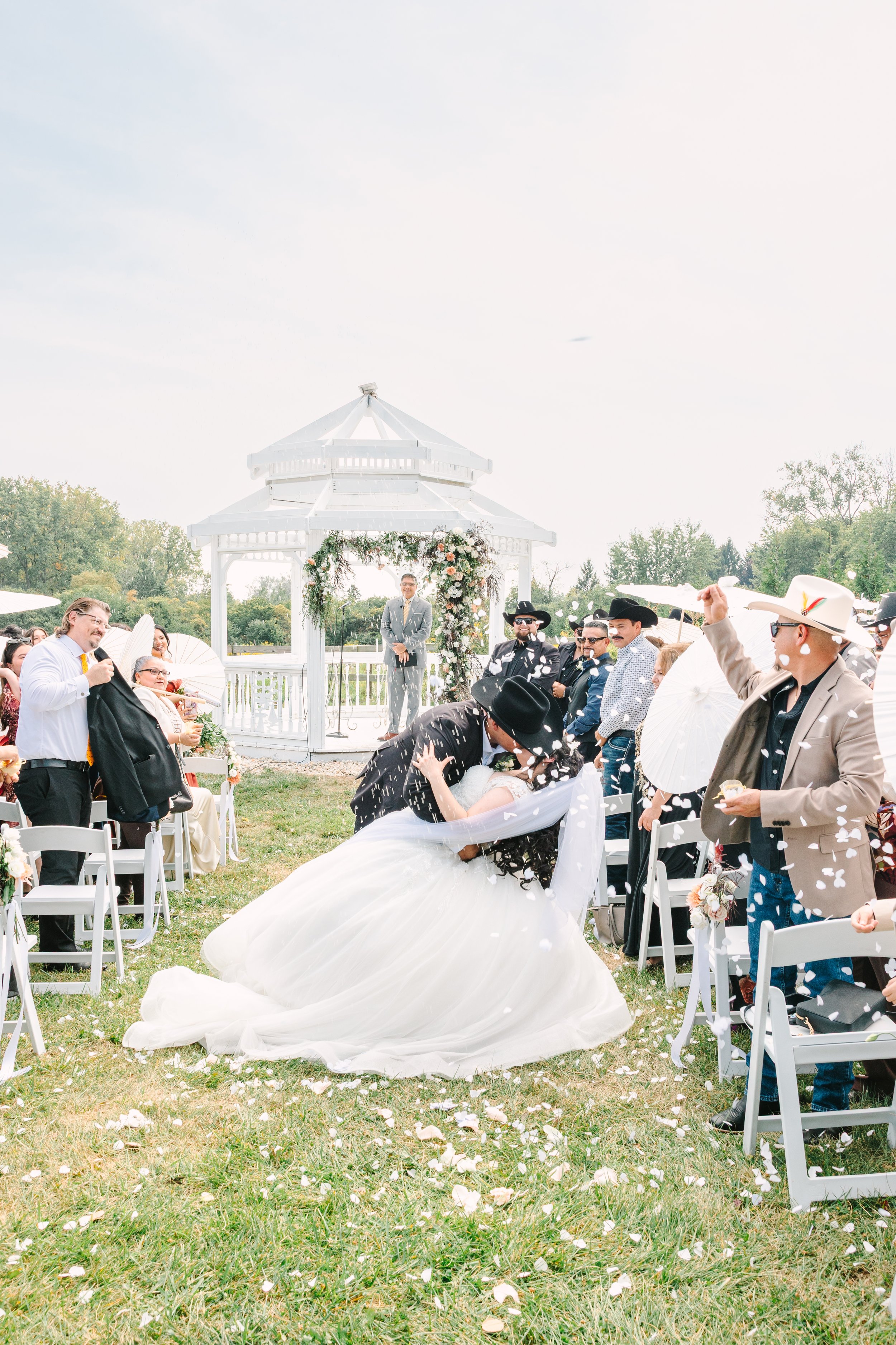 A wedding taking place outdoors under a white gazebo, with the bride and groom kissing while confetti is thrown in the air, and guests watching and celebrating.