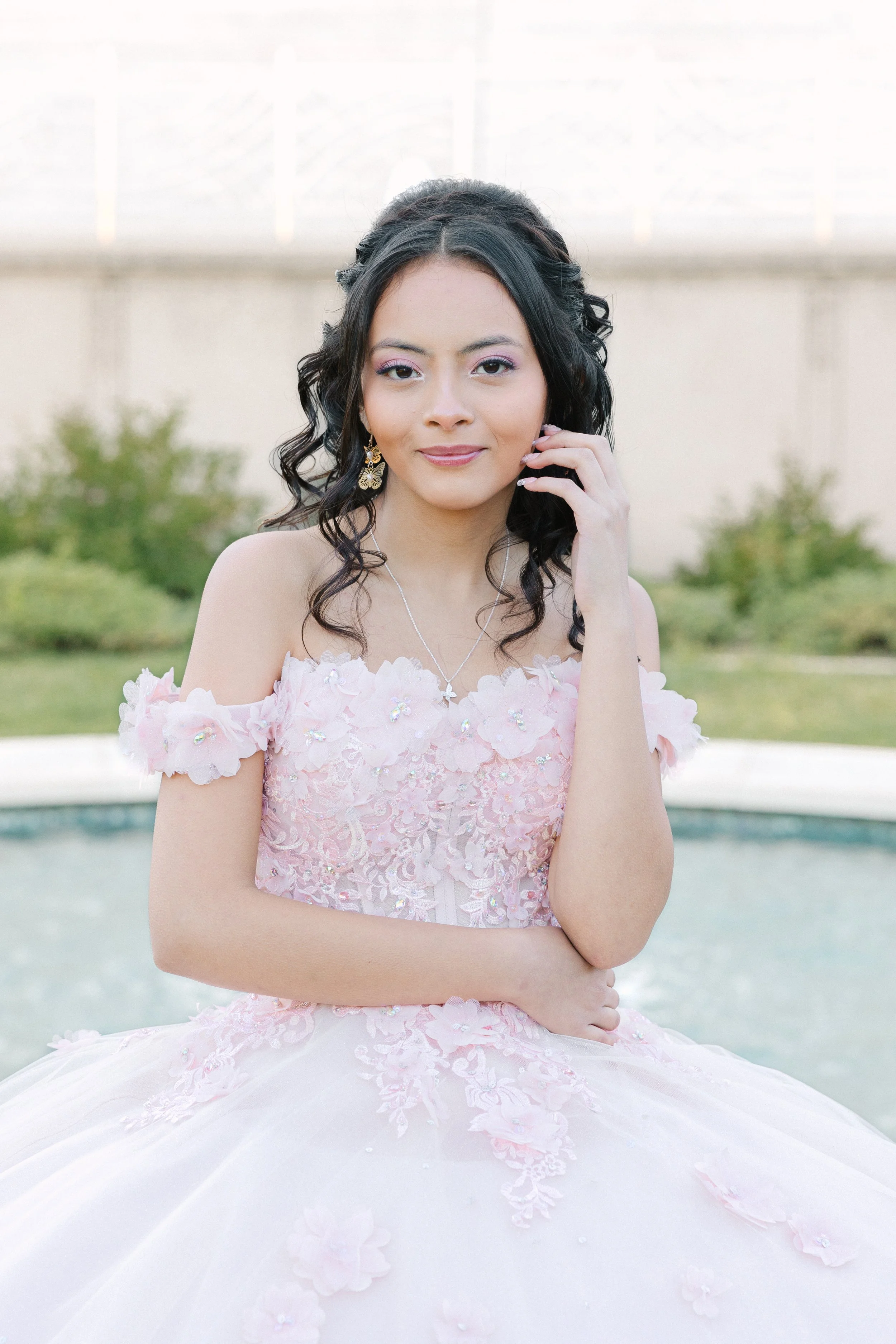 Young woman in a pink, floral wedding dress with off-shoulder sleeves, posing outdoors near a pool with greenery and a concrete wall in the background.