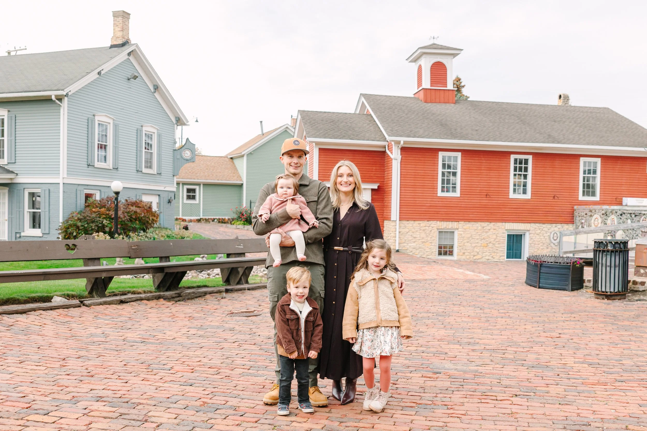 A family of five standing on a brick pathway in a colorful neighborhood with pastel-colored houses and a red building in the background.
