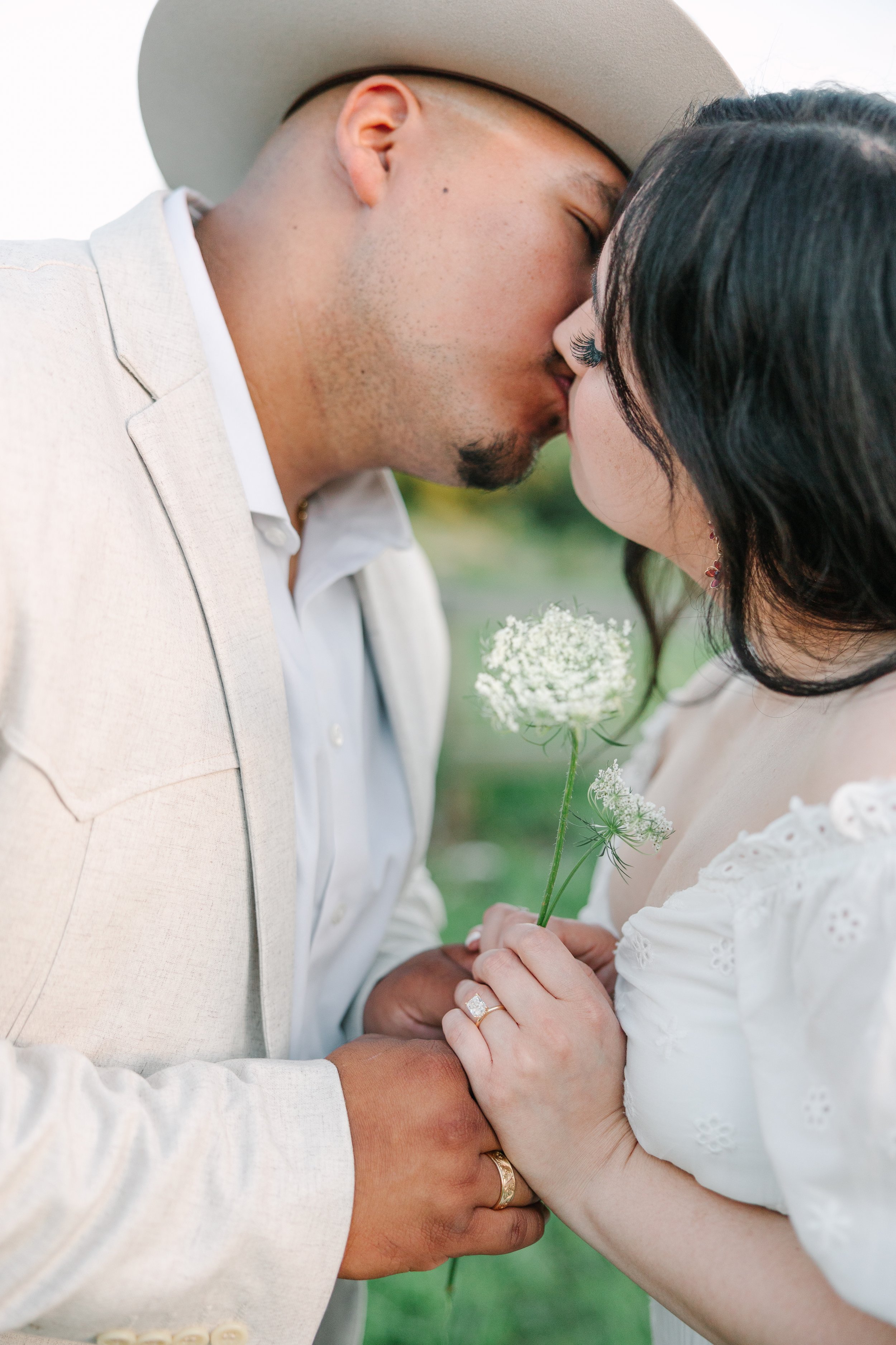 A couple sharing a kiss outdoors, the man wearing a light-colored suit and hat, holding a small white flower, with wedding rings visible on their hands.