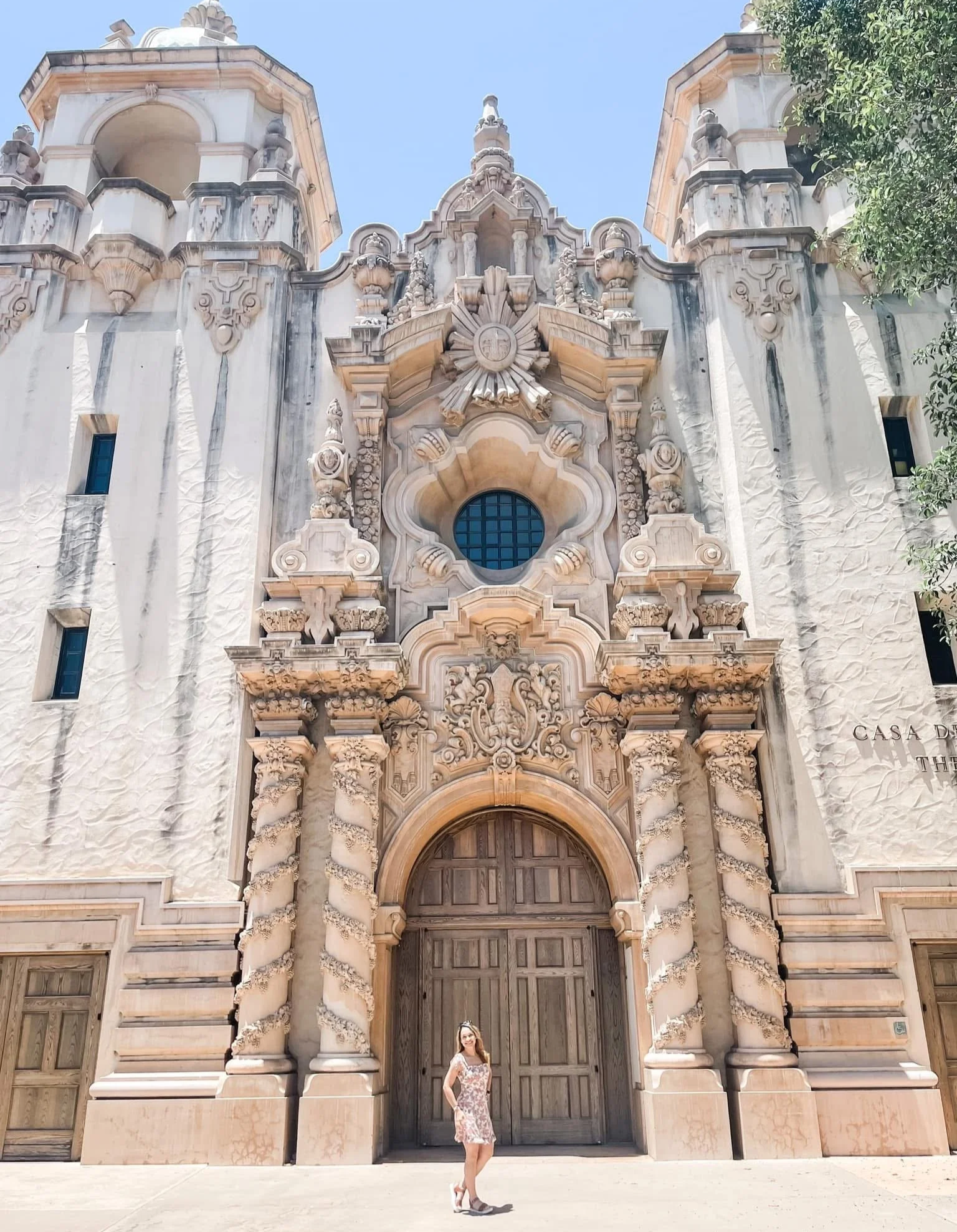 A woman in a floral dress and high heels standing in front of an ornate, historic building with intricate stonework, tall columns with spiral patterns, and a large wooden door.