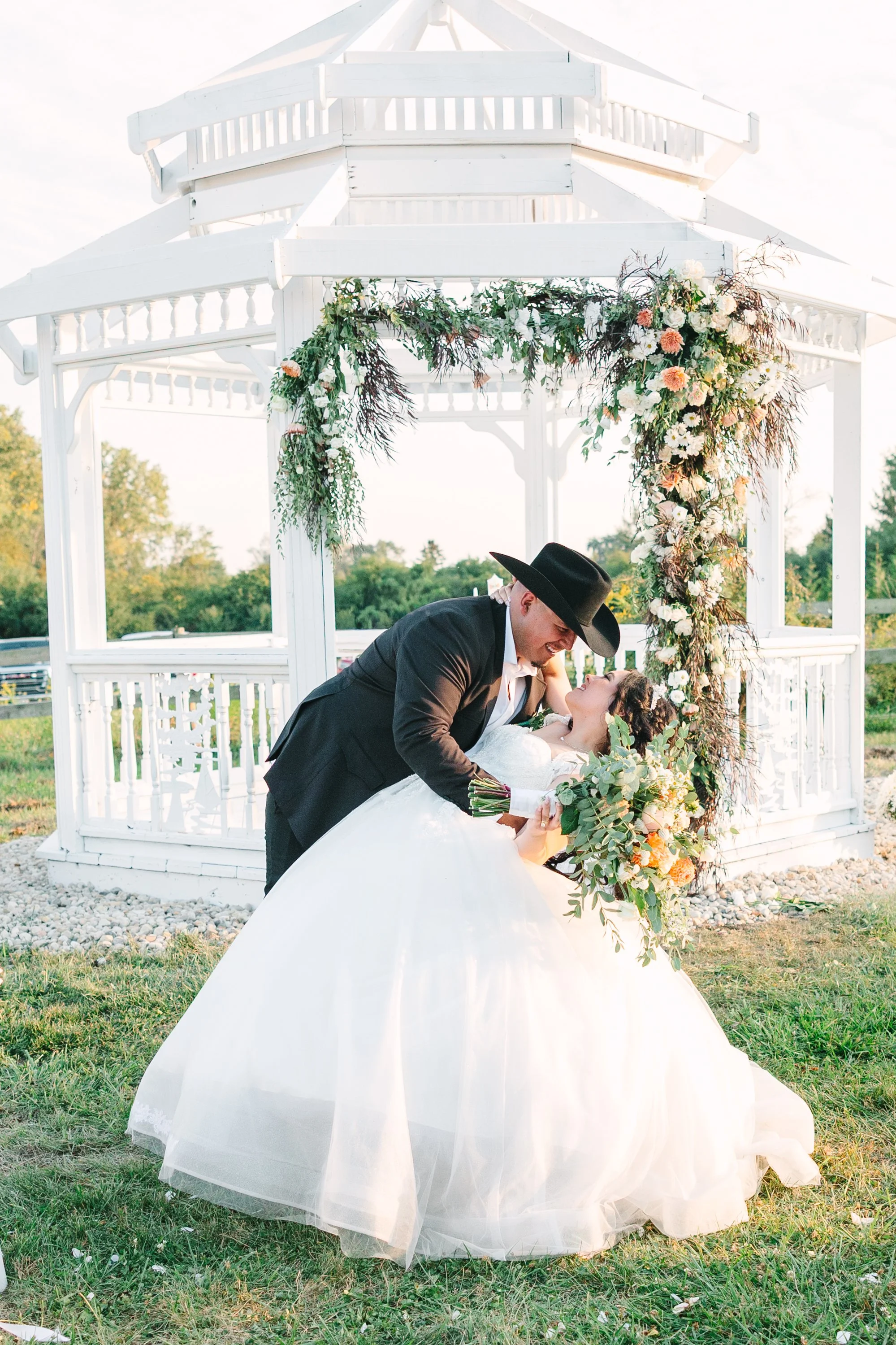 A newlywed couple sharing a romantic moment outdoors in front of a white gazebo decorated with flowers during a wedding.