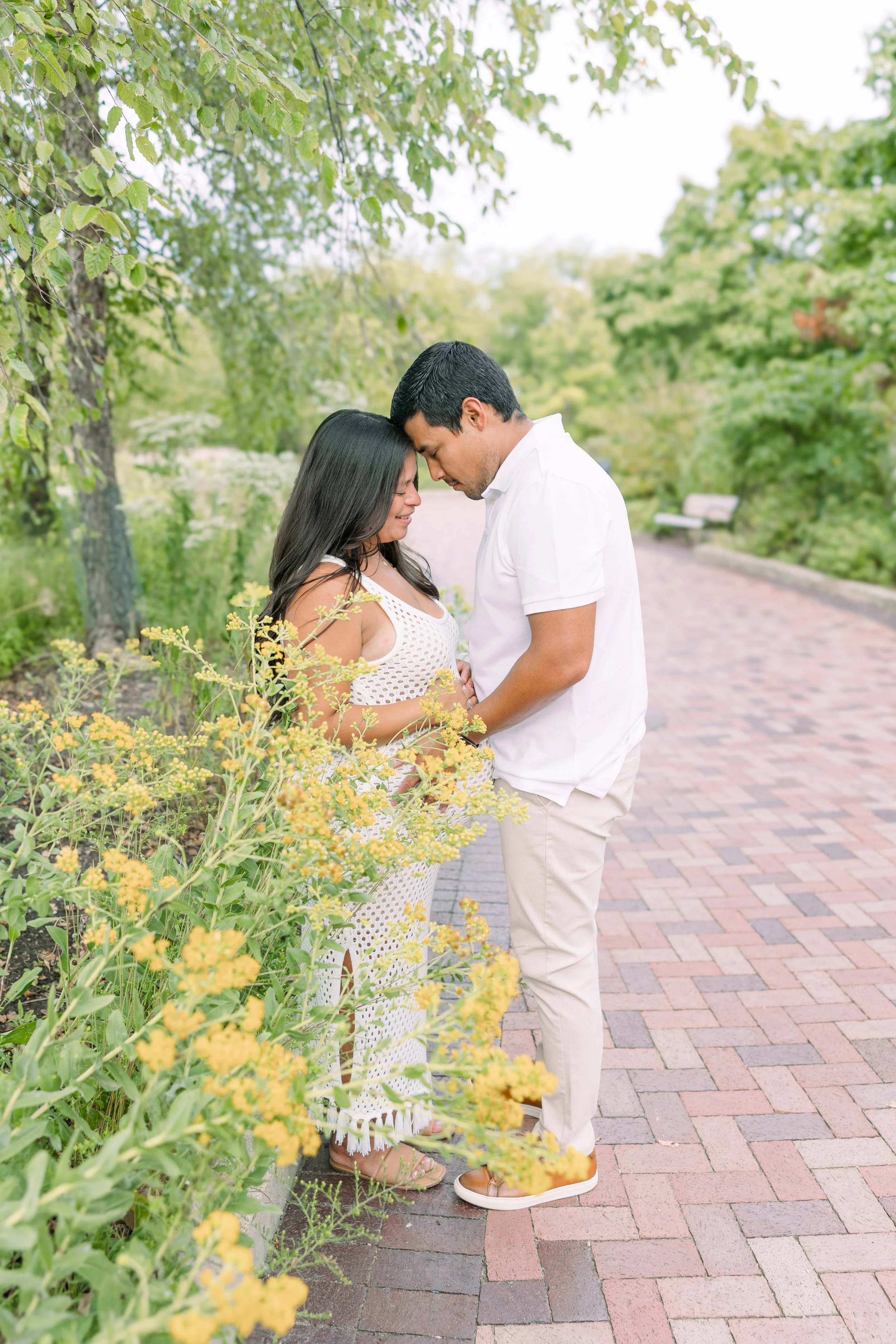 A couple standing close together on a brick pathway in a park with greenery and yellow flowers, with their foreheads touching and eyes closed.