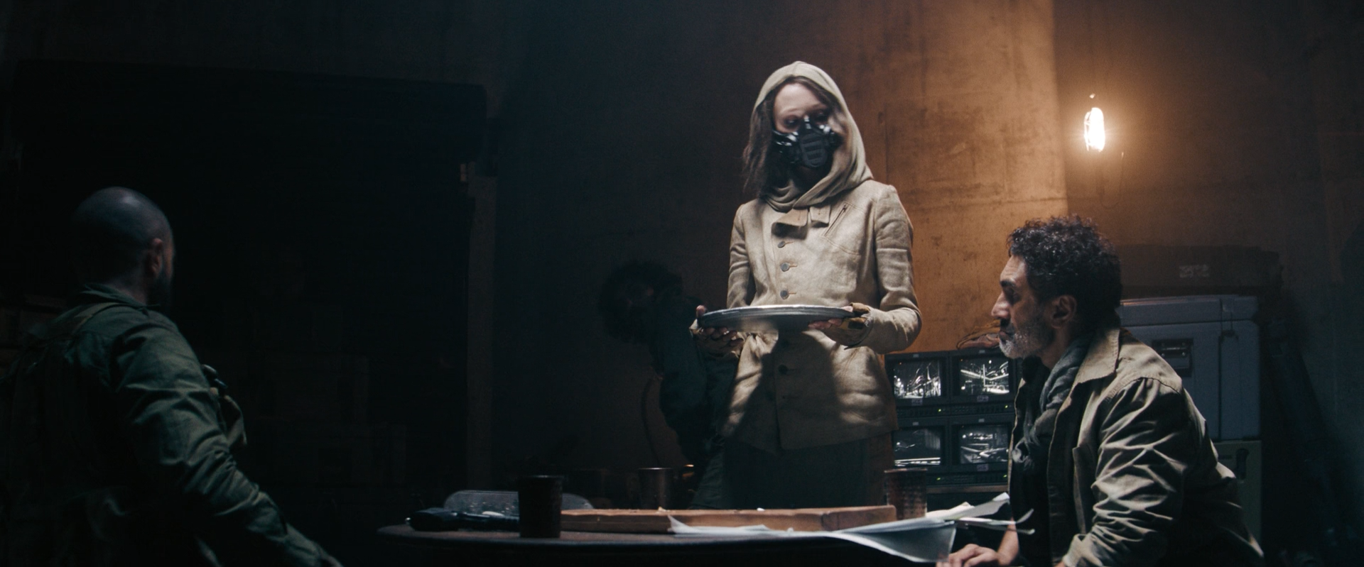 A woman wearing a beige jacket and black mask standing at a table, holding a tray, with two men seated, in a dimly lit room with surveillance screens in the background.