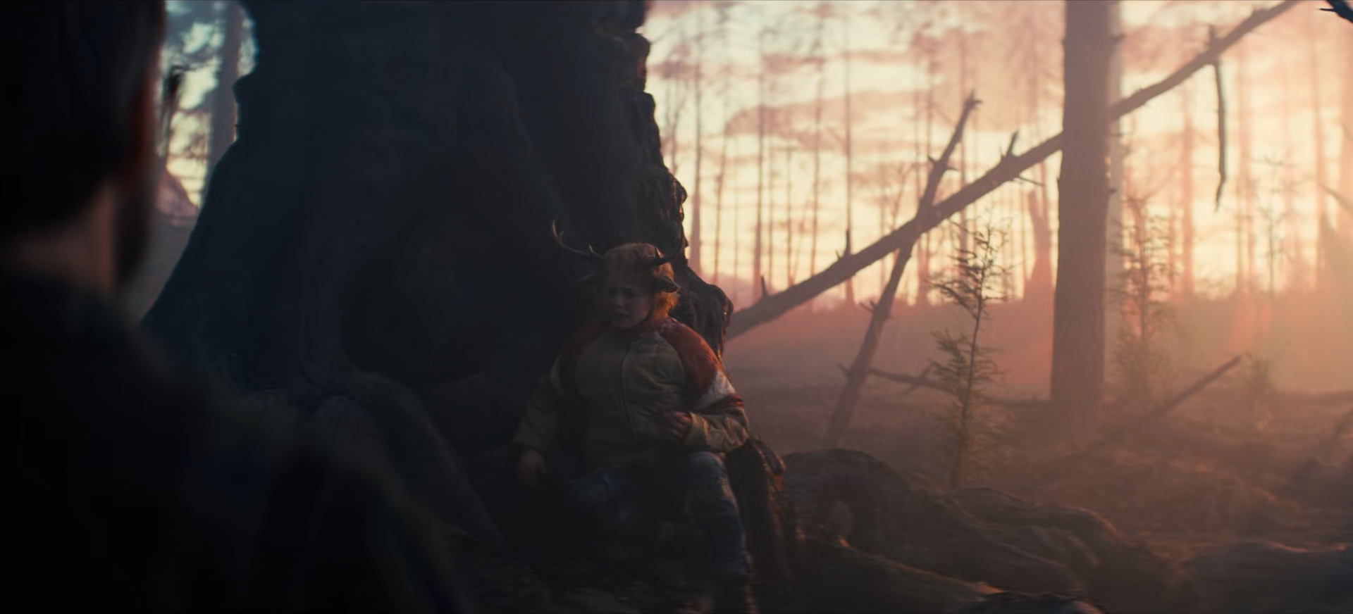 A person and a young girl sitting on a rocky outcrop near a large tree in a forest at sunset, with sunlight filtering through the trees.