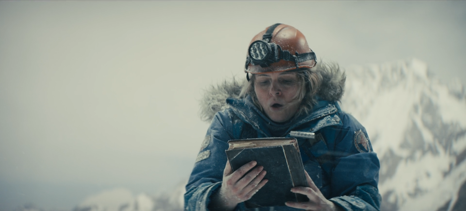 Woman in cold weather gear, including a helmet with a headlamp, looks shocked as she reads a book outdoors in a snowy landscape.