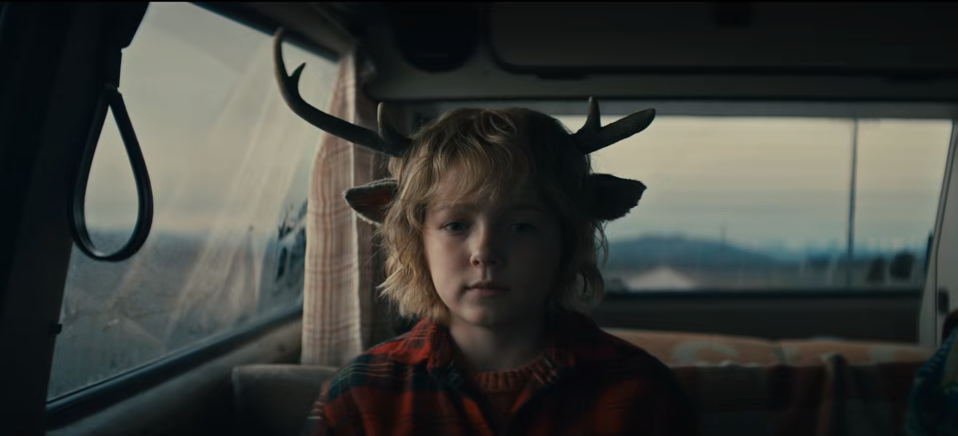 A young boy with curly hair wearing a deer headband, sitting in a vehicle with a window showing an outdoor landscape.