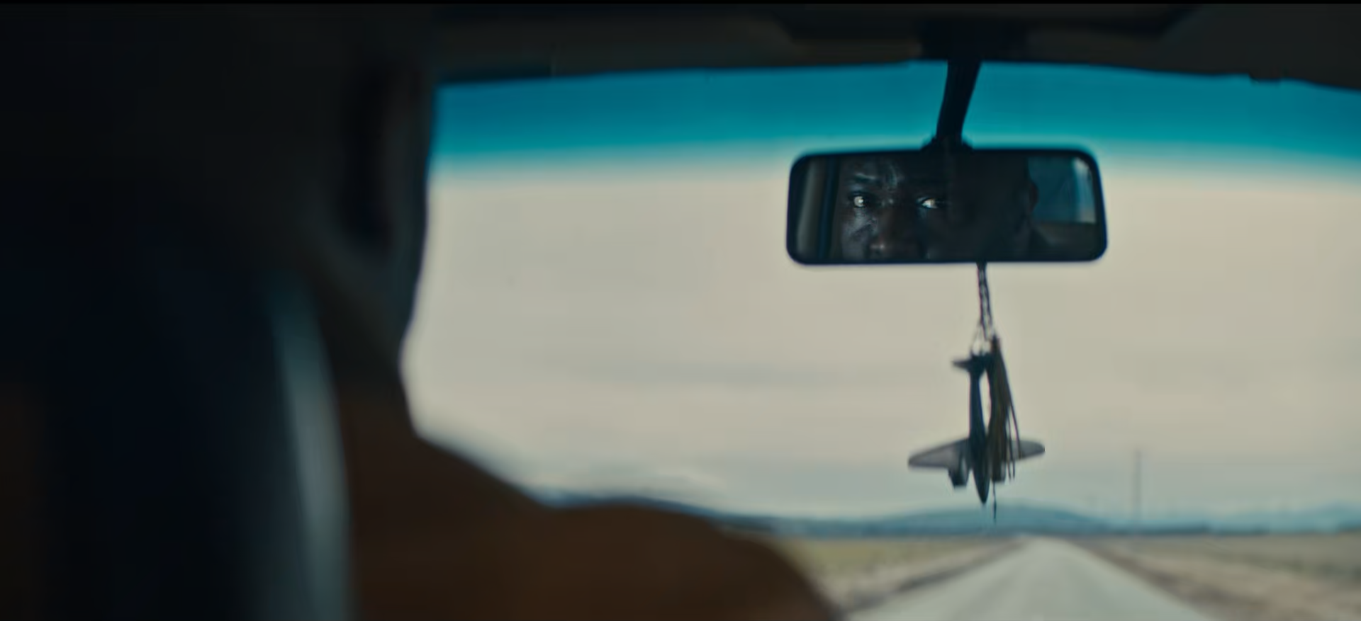 A person driving a vehicle on an open road with a clear sky, seen from behind through the windshield, with a view of the driver's face reflected in the rearview mirror.