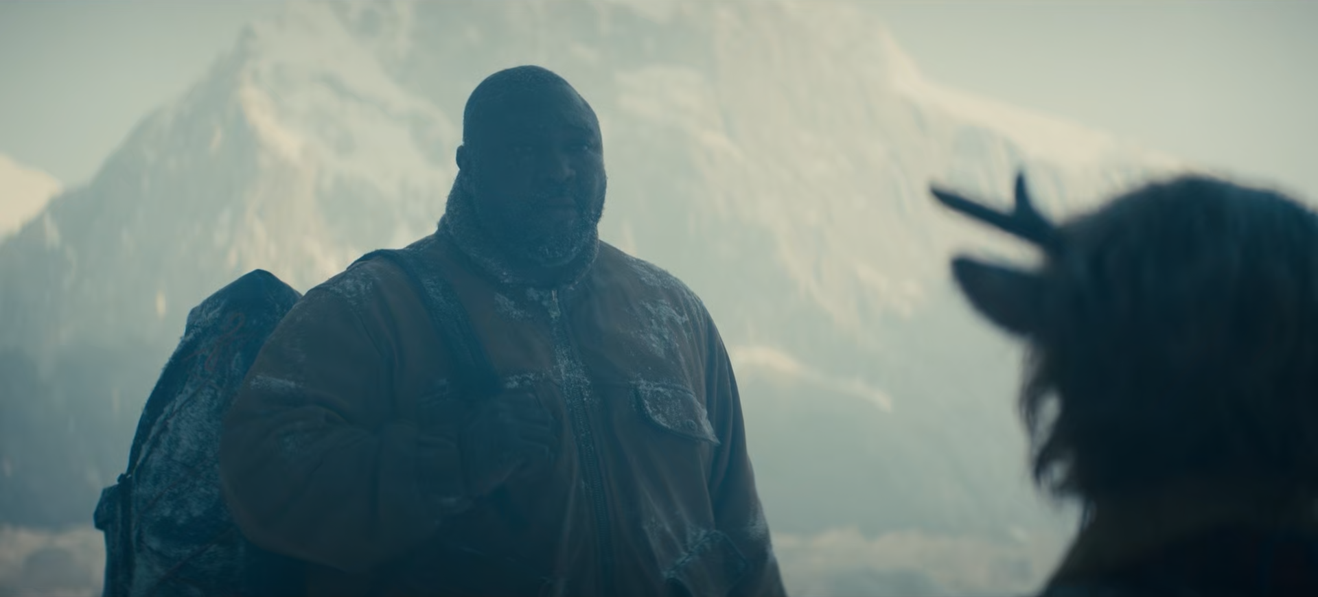 A man with a beard wearing a winter coat and backpack standing in snowy mountains, facing a yak with prominent horns in a cold, misty landscape.