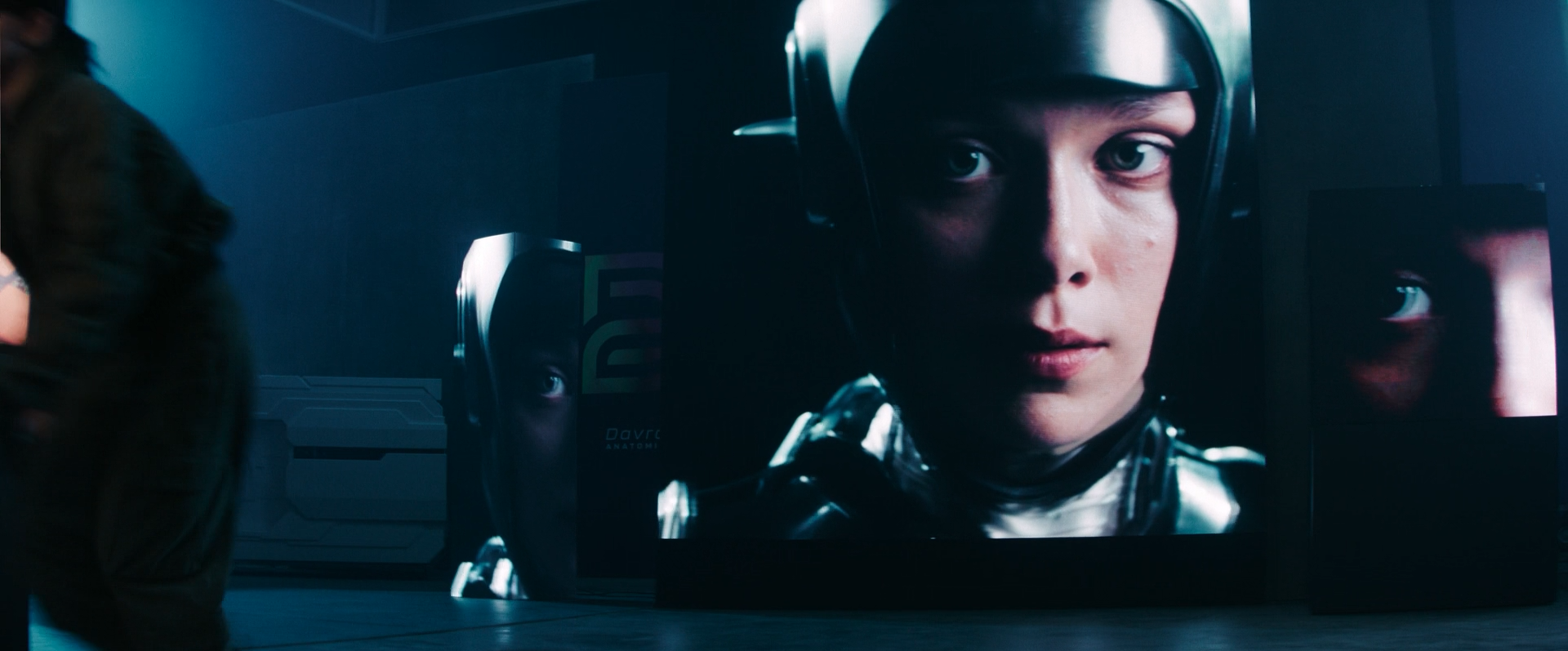 Large digital screen showing a close-up of a young woman in a futuristic helmet, with a serious expression, in a sci-fi setting with dark lighting.