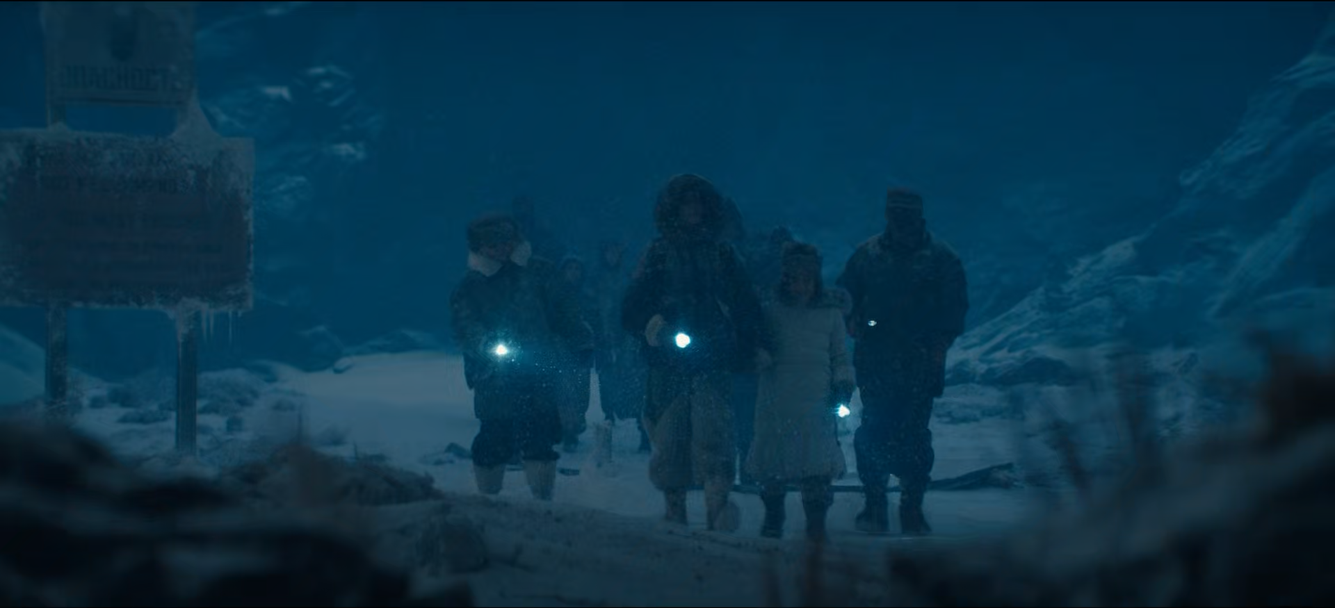 Group of people walking outside in snowy, icy environment at night, some holding flashlights, with ice-covered structures in the background.