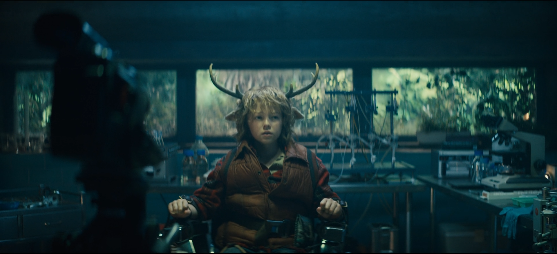 A young boy dressed as a deer with antlers and a brown vest, sitting in a laboratory surrounded by scientific equipment, with a window showing greenery in the background.