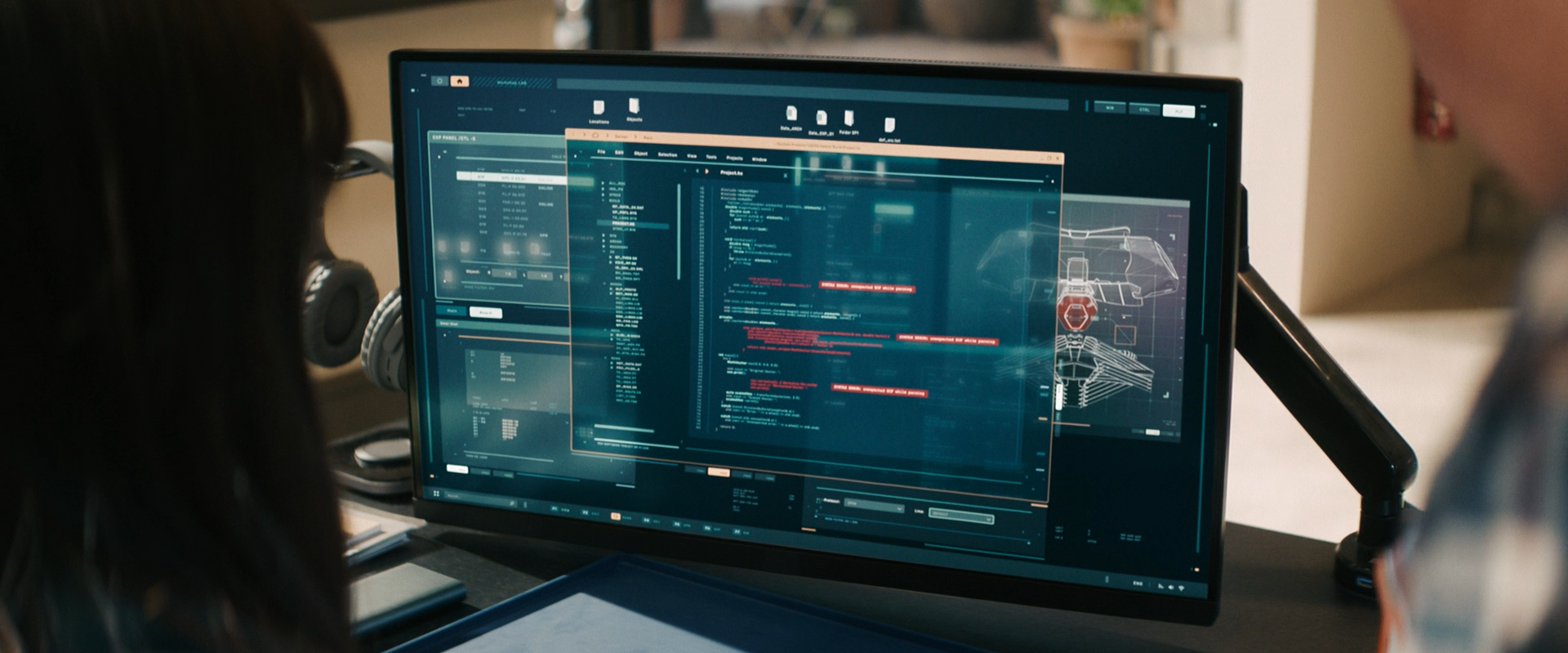 Computer monitor displaying a blue-themed software interface with code, diagrams, and data, with people working nearby.