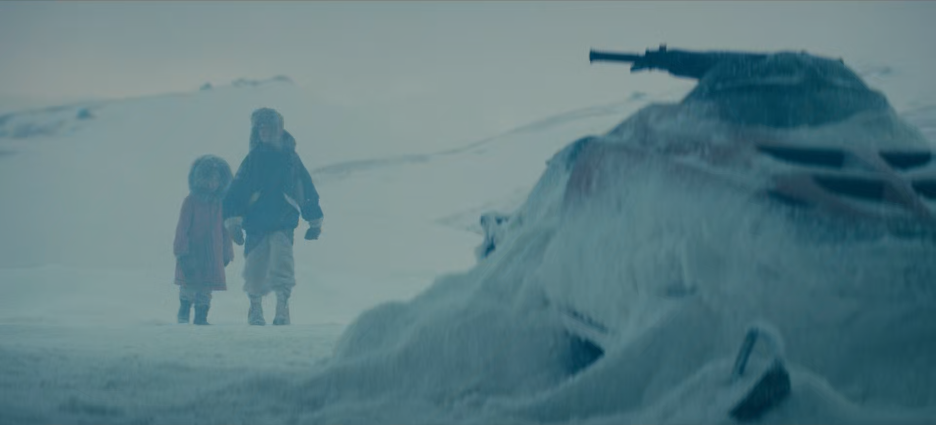 Two people wearing winter clothing walking in a snow-covered landscape near a large piece of machinery or debris.