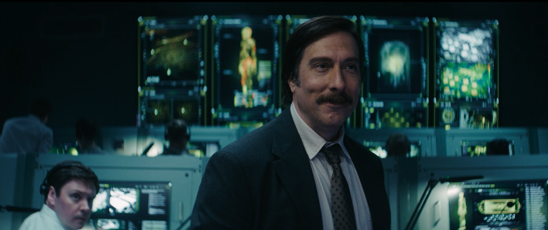 Scene from a control room with multiple monitors displaying digital data, with a man with dark hair and a mustache wearing a suit in the foreground, smiling. Others are working at computers in the background.
