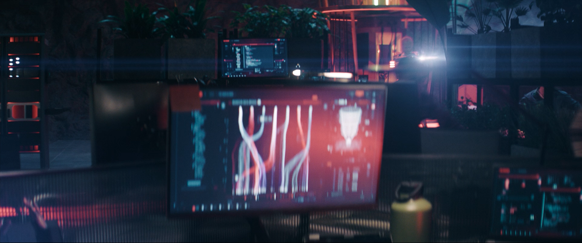 A dimly lit room with computer monitors displaying red and blue lines and graphics, pink and purple lighting, and plants in the background, creating a cyberpunk or futuristic atmosphere.