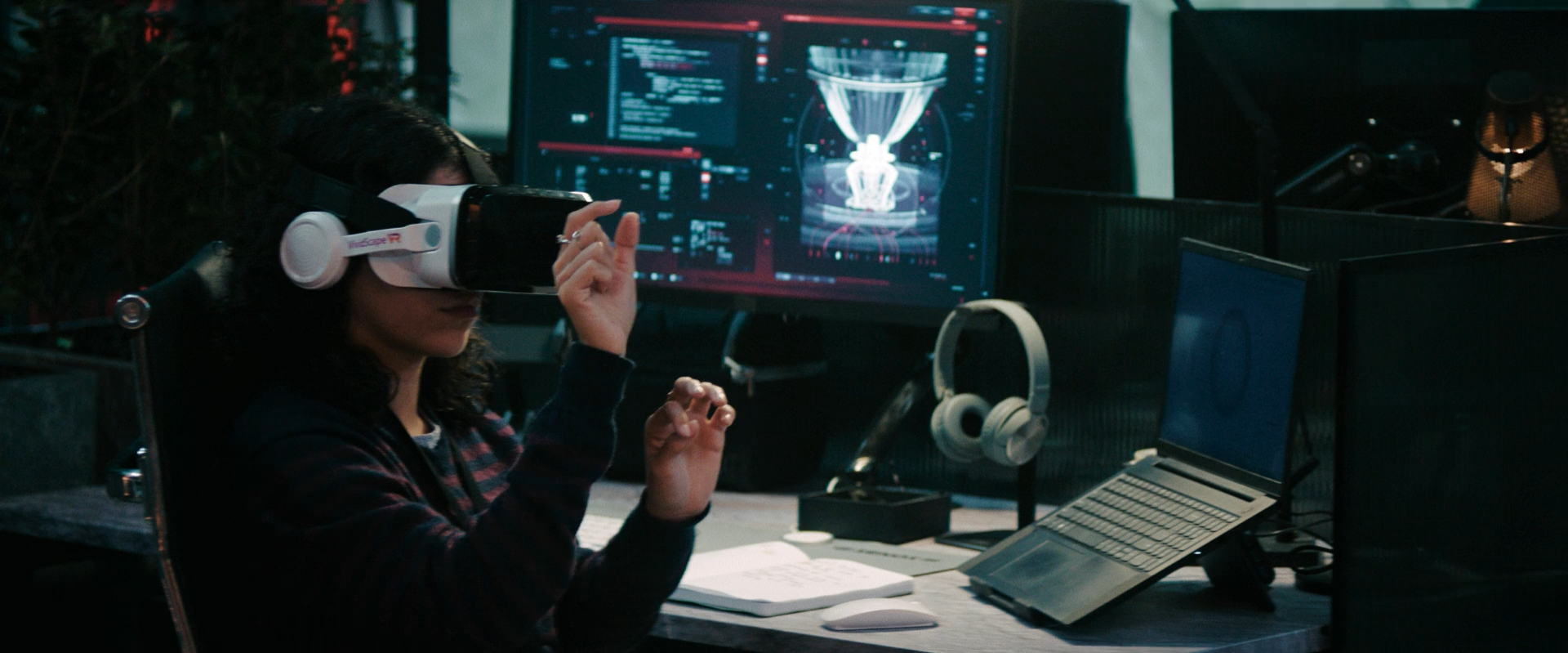 A woman wearing a virtual reality headset sitting at a desk with a large monitor, a laptop, and headphones, in a dark room.