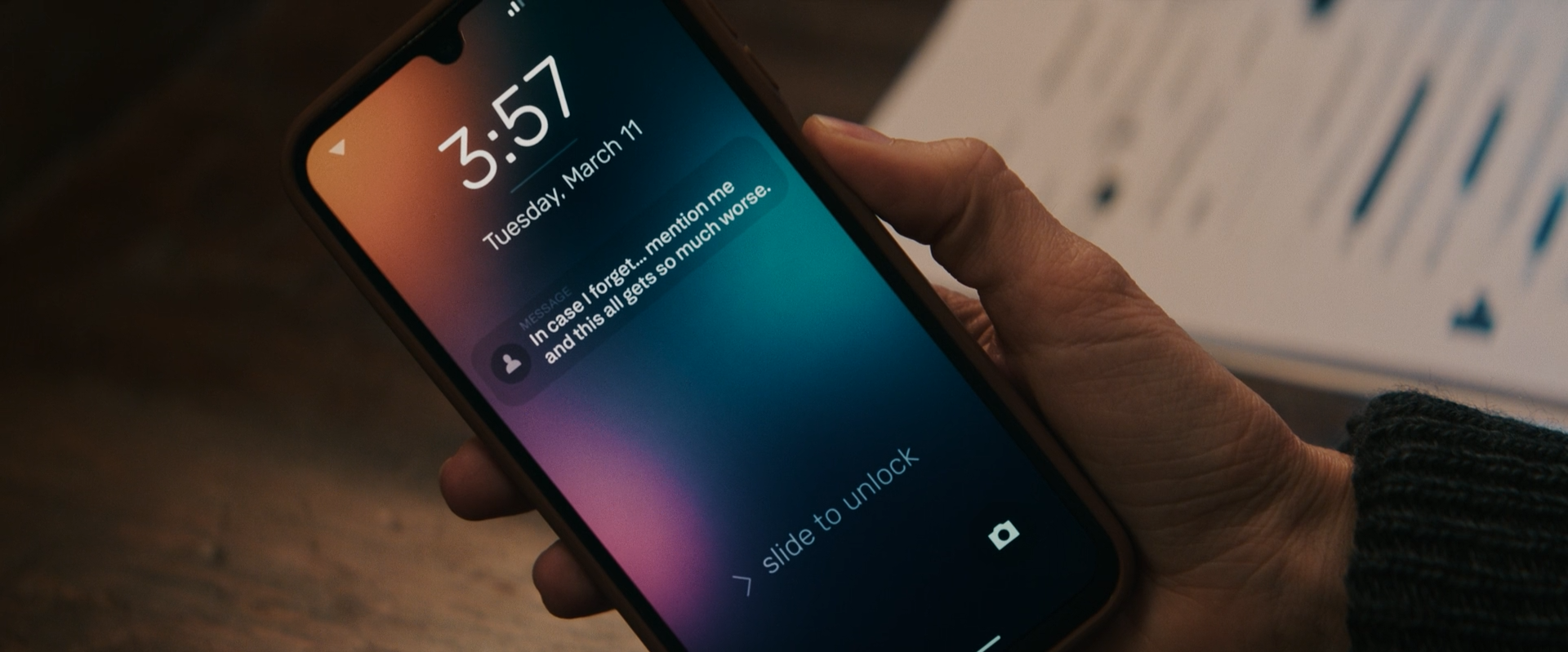 A smartphone displaying a lock screen with a message notification, showing the time as 3:57 and the date as Tuesday, March 11. The message says, 'In case I forget... mention me and this all gets so much worse.' The phone is being held in someone's ha