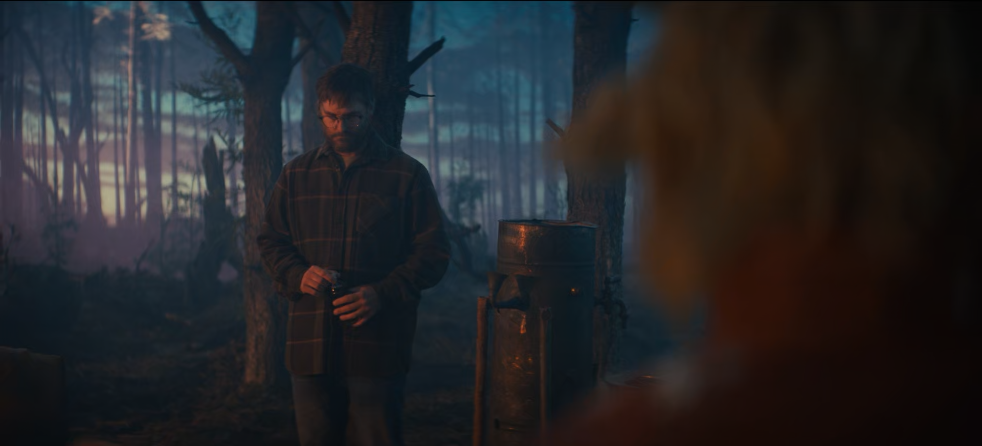 A man with glasses and a beard standing in a dimly lit forest at dusk, holding a small object in his hands near a tree.